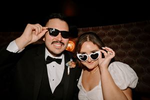Bride and groom in sunglasses posing playfully at wedding reception, groom in tuxedo, bride in white dress.