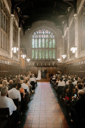 Wedding ceremony in grand Gothic hall with stained glass window, bride and groom at altar, guests seated in wooden pews.