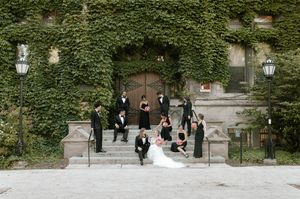 Wedding party in black attire poses on ivy-covered stone building steps with bride and groom centered.