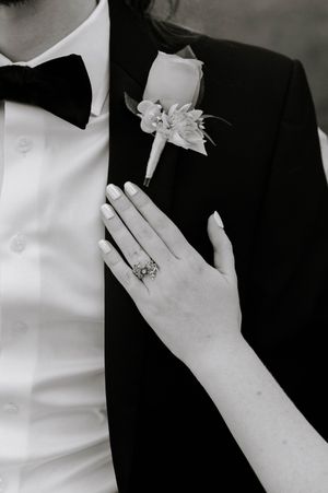 Black and white wedding photo of bride's hand with ring resting on groom's tuxedo lapel with boutonniere.