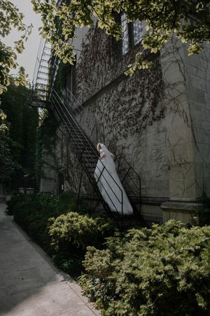 Bride in white gown standing on iron fire escape stairs of ivy-covered stone church building surrounded by lush green shrubs.