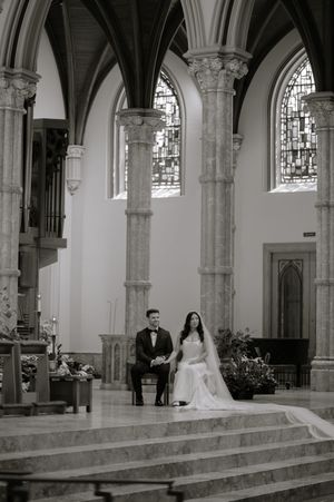 Black and white photo of bride and groom seated at church altar with gothic arches and stained glass windows.