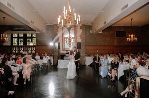 Bride and groom share first dance in elegant ballroom with chandelier and seated wedding guests.