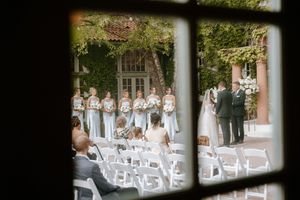 Outdoor wedding ceremony viewed through window with bridesmaids in blue gowns and guests seated in white chairs.