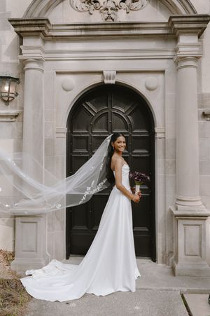 Bride in white gown with flowing veil stands before ornate stone archway and black door holding bouquet.