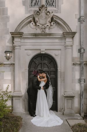 Bride and groom share a kiss in front of an elegant stone arched doorway with decorative carvings.