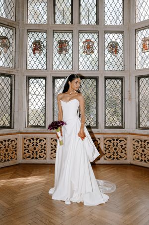 Bride in strapless white gown holding purple bouquet stands before ornate stained glass windows on herringbone wood floor.