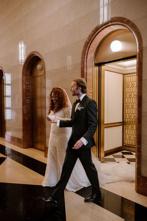 Bride and groom walking through elegant marble hallway with Art Deco elevators on their wedding day.