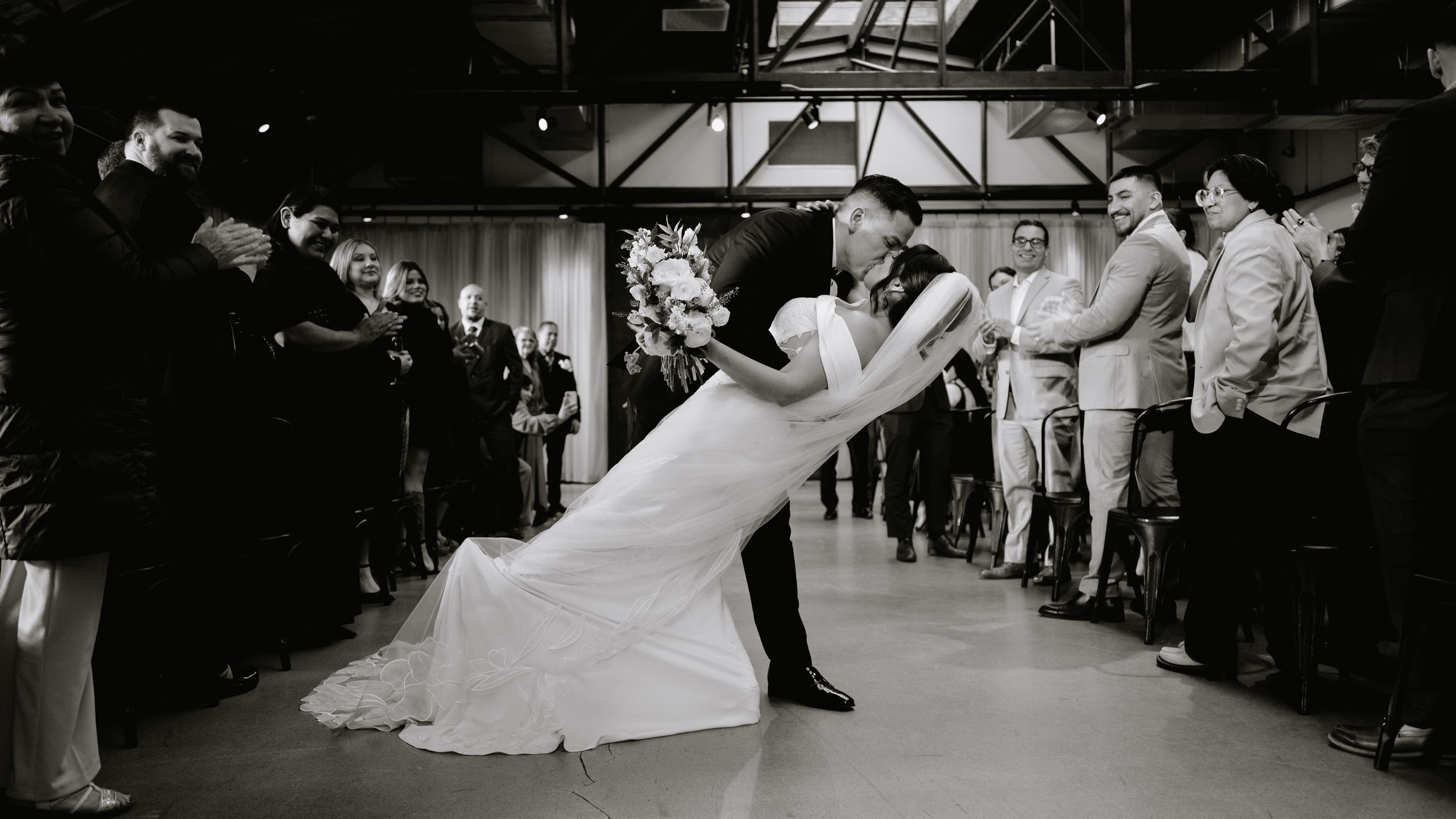 Black and white photo of bride and groom sharing a romantic dip kiss surrounded by wedding guests.