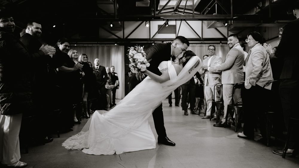 Black and white photo of bride and groom sharing a romantic dip kiss surrounded by wedding guests.