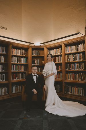 Couple in wedding attire posing in a library, groom in tuxedo seated beside bride in white gown holding bouquet.