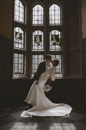 Bride and groom share a romantic dip kiss in front of stunning stained glass windows in an elegant wedding venue.