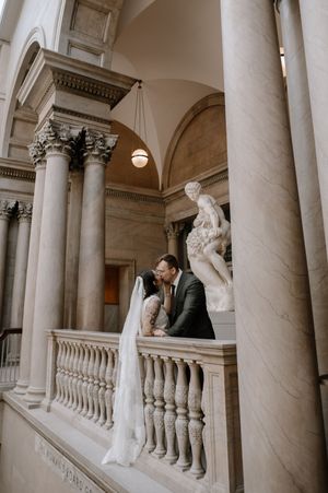 Bride and groom share a kiss on a marble balcony beside a classical statue in an elegant columned hall.