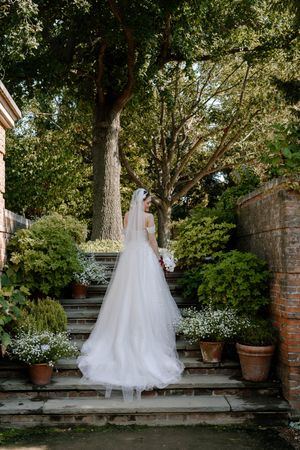 Bride in white gown and veil stands on garden steps surrounded by lush greenery and potted flowers.