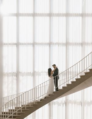 A bride and groom share a romantic embrace on an elegant staircase before a grand floor-to-ceiling sheer curtain backdrop.