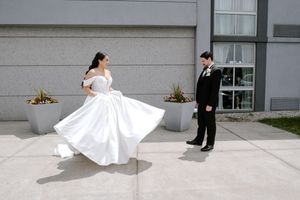 Bride twirling in white wedding gown while groom watches outdoors near modern building on sunny day.