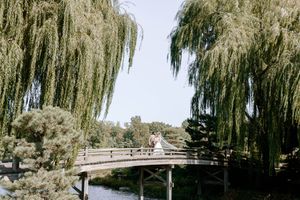 Couple on wooden bridge framed by weeping willows in a serene Japanese garden on a sunny day.