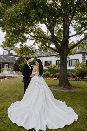 Bride in a flowing white gown and groom in a black tuxedo embrace under a large tree on a lush green lawn.