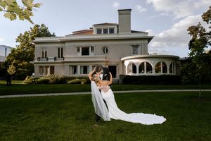 Bride and groom share romantic dip kiss on lush lawn before elegant mansion wedding venue on sunny day.