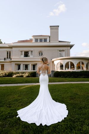 Bride in elegant backless white mermaid gown with lace detail and long train stands before a grand estate.