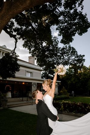 Groom lifting bride in white gown outdoors, she raises bouquet joyfully, elegant manor and large tree in background.