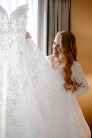 Smiling bride with long red hair holding up her white lace wedding dress near a window before her ceremony.