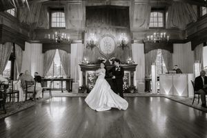 Black and white photo of bride and groom sharing first dance in elegant ballroom with chandeliers and fireplace.