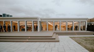 Modern glass-walled event venue with guests visible inside, featuring wide steps and paved courtyard under overcast sky.