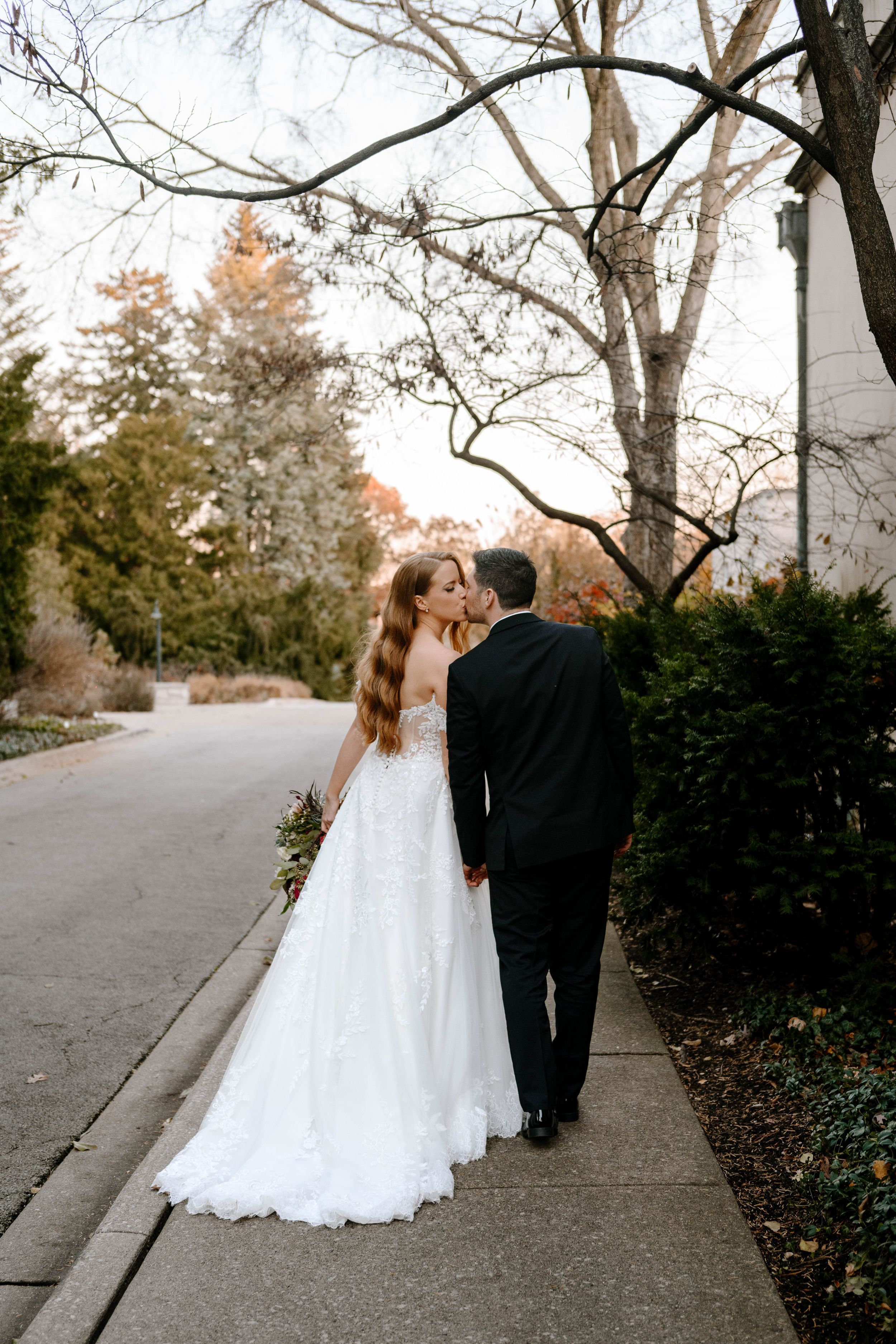 Bride in white lace gown and groom in black suit share a kiss while walking on a sidewalk surrounded by autumn trees.