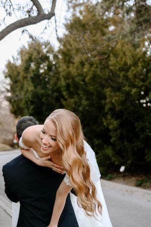Smiling bride in white lace dress embracing groom in black suit outdoors with lush green trees in background.