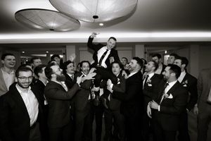 Groomsmen joyfully toss groom into the air during black and white wedding celebration photo.