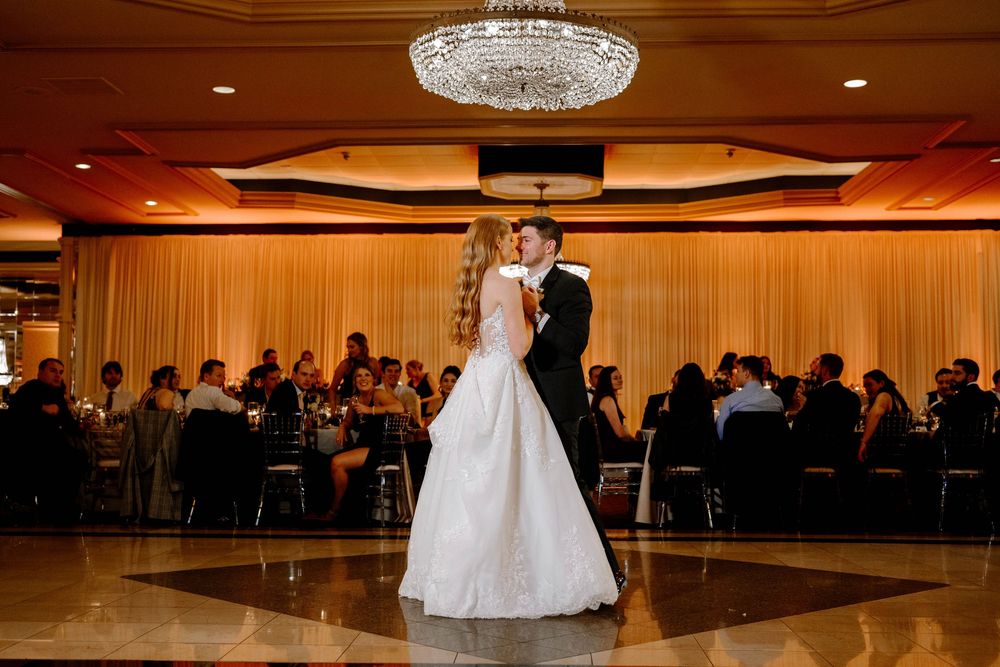 Bride and groom share first dance under crystal chandelier at elegant wedding reception with guests seated at tables.