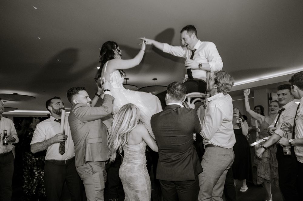 Black and white wedding photo of bride and groom being lifted by guests during lively reception dance celebration.