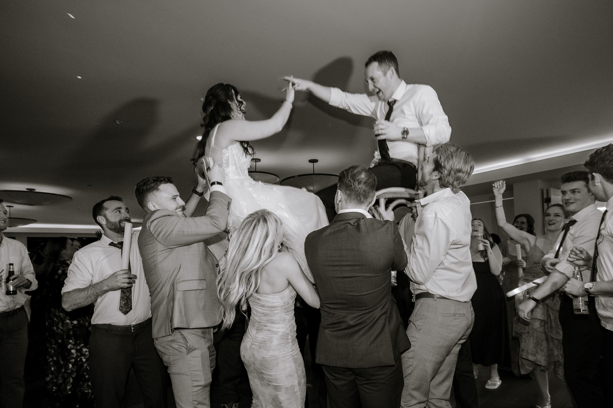 Black and white wedding photo of bride and groom being lifted by guests during lively reception dance celebration.