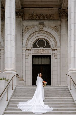 Bride in a long white gown with cathedral train stands on steps of ornate building holding colorful bouquet.