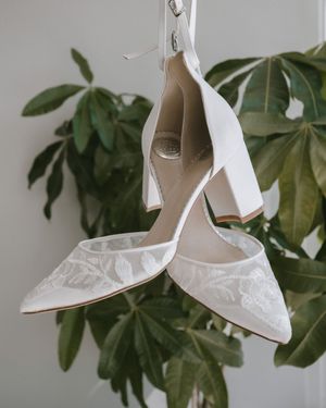 White lace block heel bridal shoes hanging against green tropical leaves backdrop.