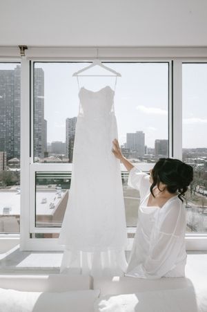 Bride in white robe admiring her wedding dress hanging by a large window with city skyline views.