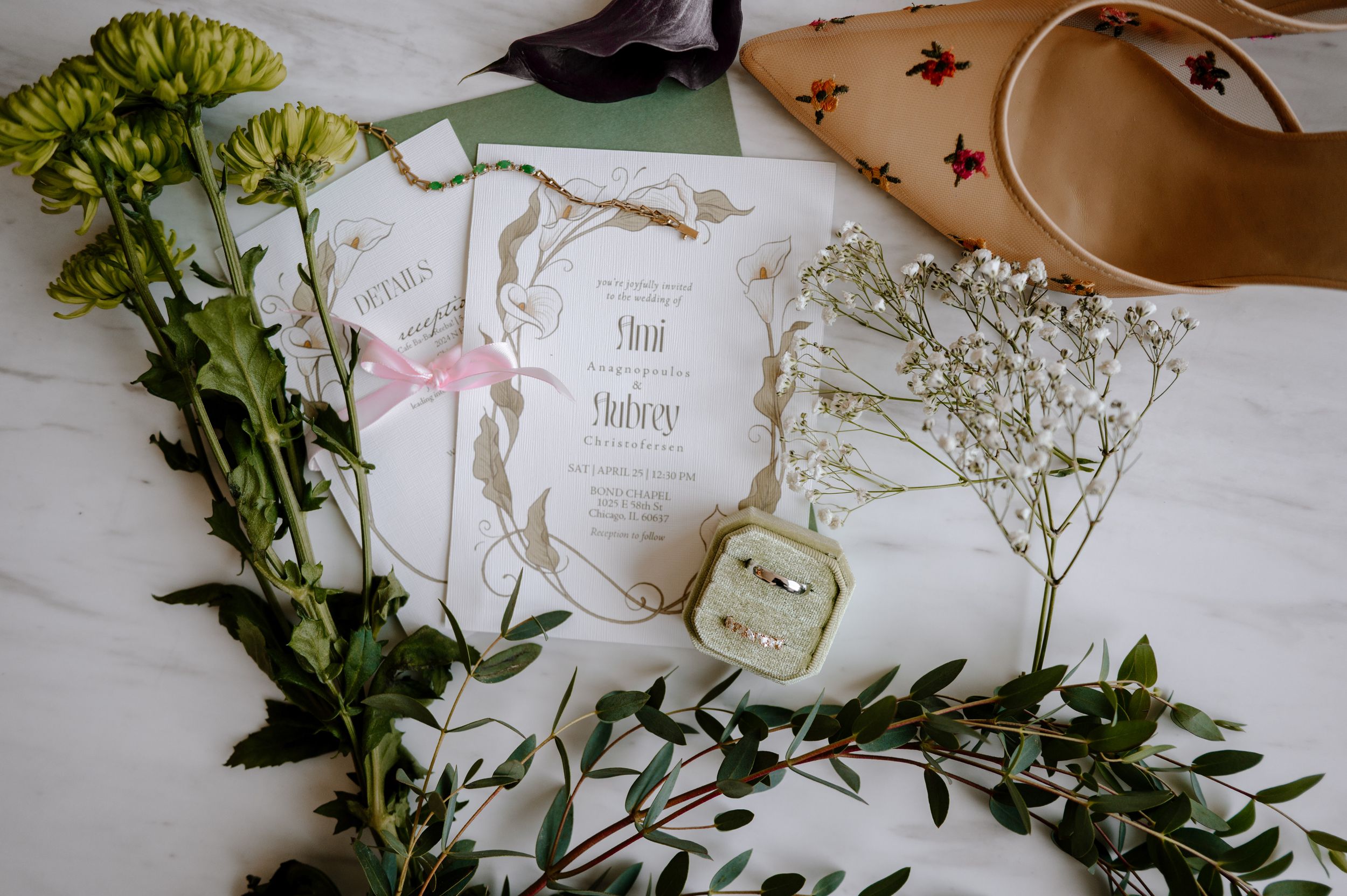 Wedding flat lay with floral invitation suite, ring box, baby's breath, greenery, and tan heels on marble surface.