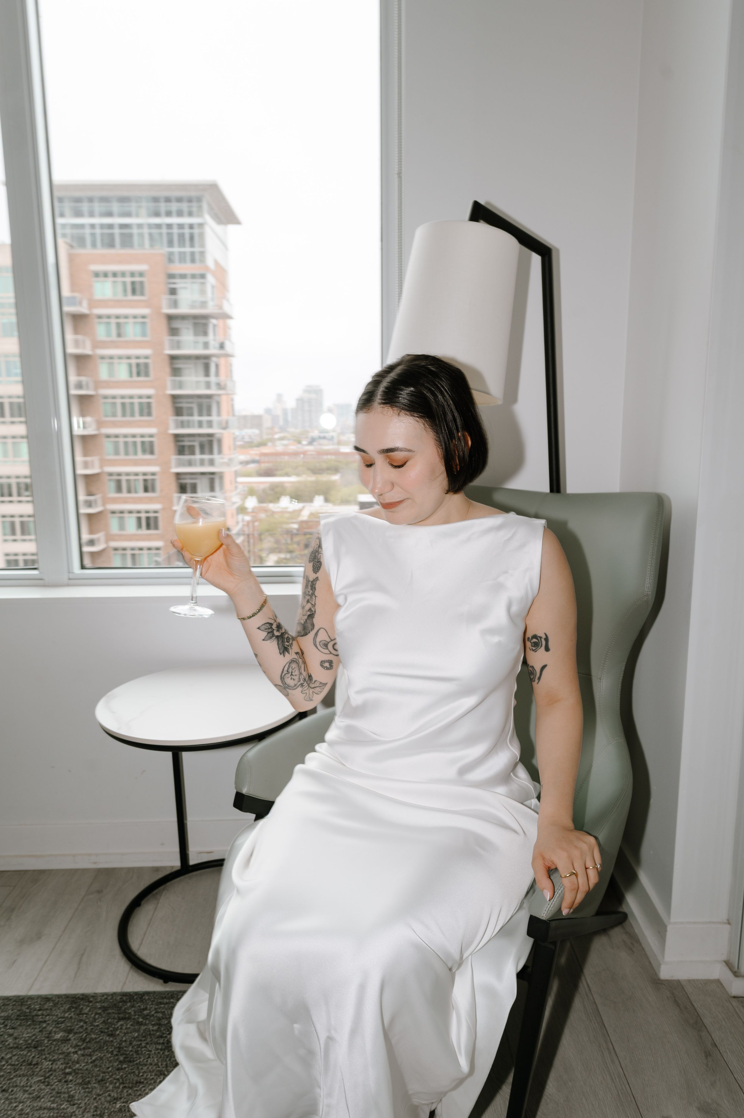Tattooed bride in white satin dress sits in chair holding champagne glass near window with city view.