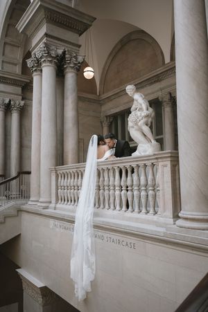 Bride and groom share a kiss on a grand marble staircase with classical columns and sculpture at an elegant wedding venue.