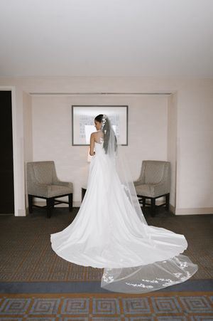 Bride in white gown with long cathedral veil stands before window in elegant hotel room on wedding day.