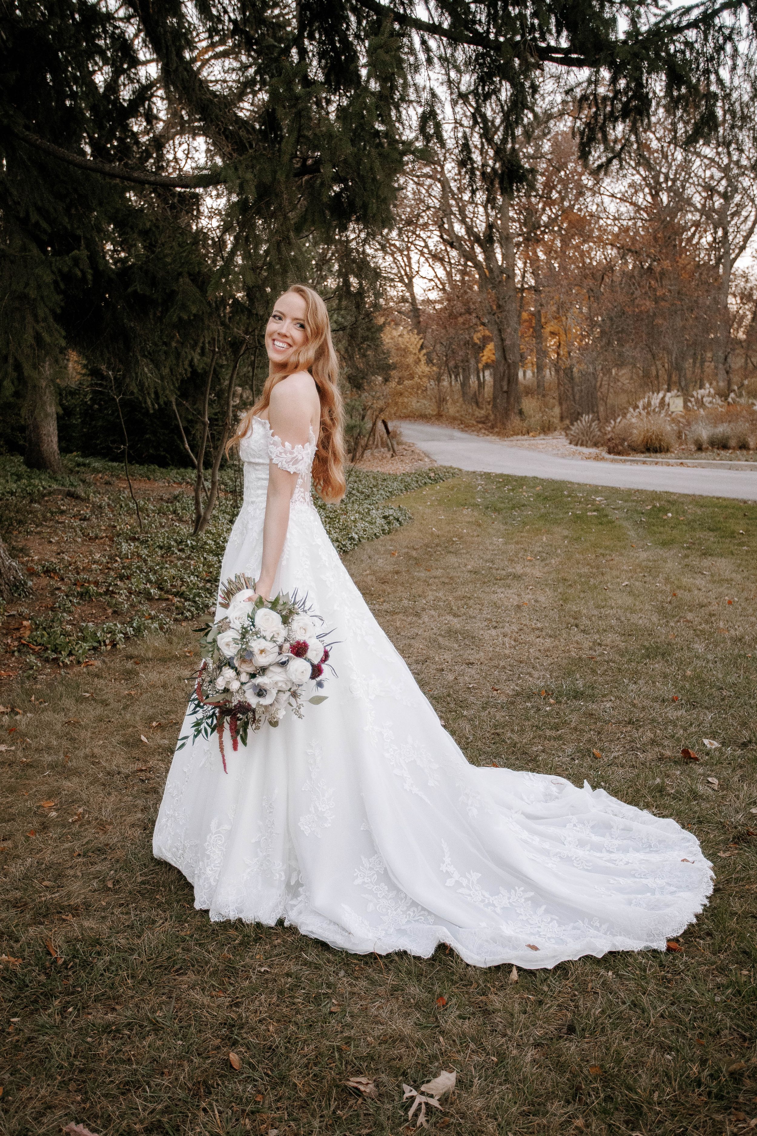 Smiling bride in a lace off-shoulder gown with a long train, holding a bouquet in an autumn park.
