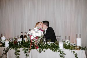 Bride and groom share a kiss at floral-adorned sweetheart table with greenery garland and white candles.