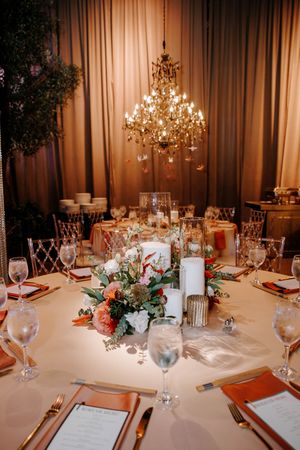 Elegant wedding reception table with floral centerpiece, candles, gold cutlery, and a chandelier in a warmly lit ballroom.