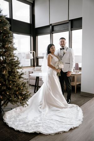 Bride in elegant white gown with long train and groom in white tuxedo jacket pose beside a decorated Christmas tree.