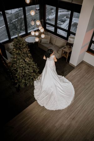 Aerial view of a bride in a stunning white wedding gown with long train standing near a decorated Christmas tree indoors.