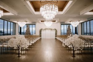Elegant wedding ceremony venue with crystal chandeliers, floral arch, white cherry blossom trees, and Christmas trees in a bright ballroom.