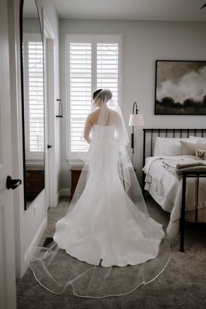Bride in white wedding gown and cathedral veil stands facing window in elegant bedroom before ceremony.