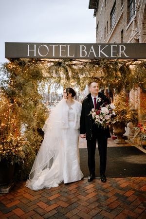 Bride and groom holding hands outside Hotel Baker, bride in white gown with veil, groom in black suit with floral bouquet.