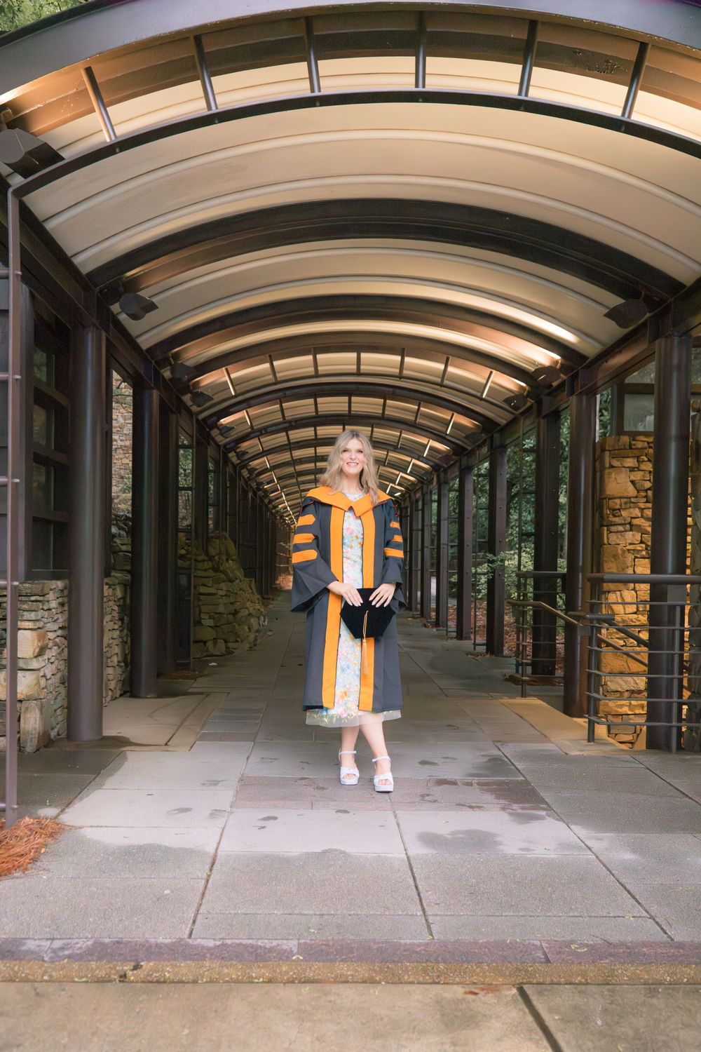 Graduate in orange and black academic regalia poses in an arched glass corridor surrounded by stone walls.